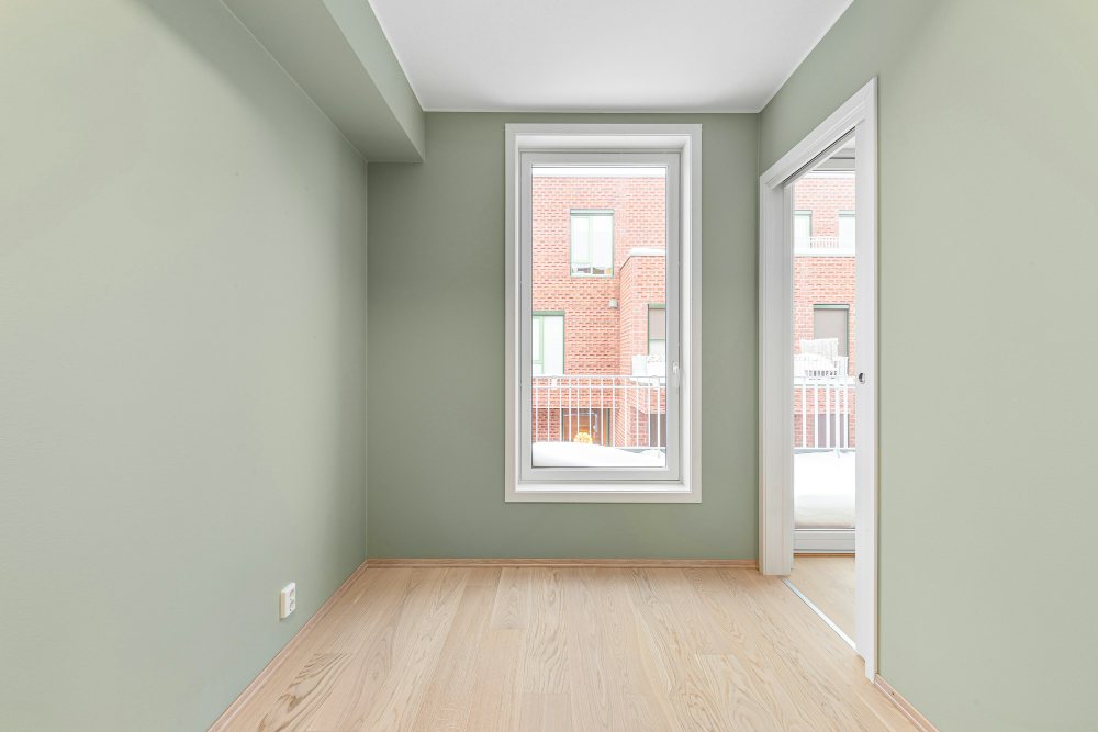 An empty room in daylight: sage green walls, a white-framed window, a white baseboard and a pale wood floor